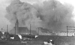 De eerste inslagen in het gedeelte achter de Schenkkade dichtbij de St. Liduinakerk gezien vanuit Voorburg over de spoorlijn Den Haag - Leiden. Foto door D. Dijkstra. Verzameling HGA, fotonr. 1945 III 63 of 64. Postbus 12600, 2500 DJ  Den Haag.